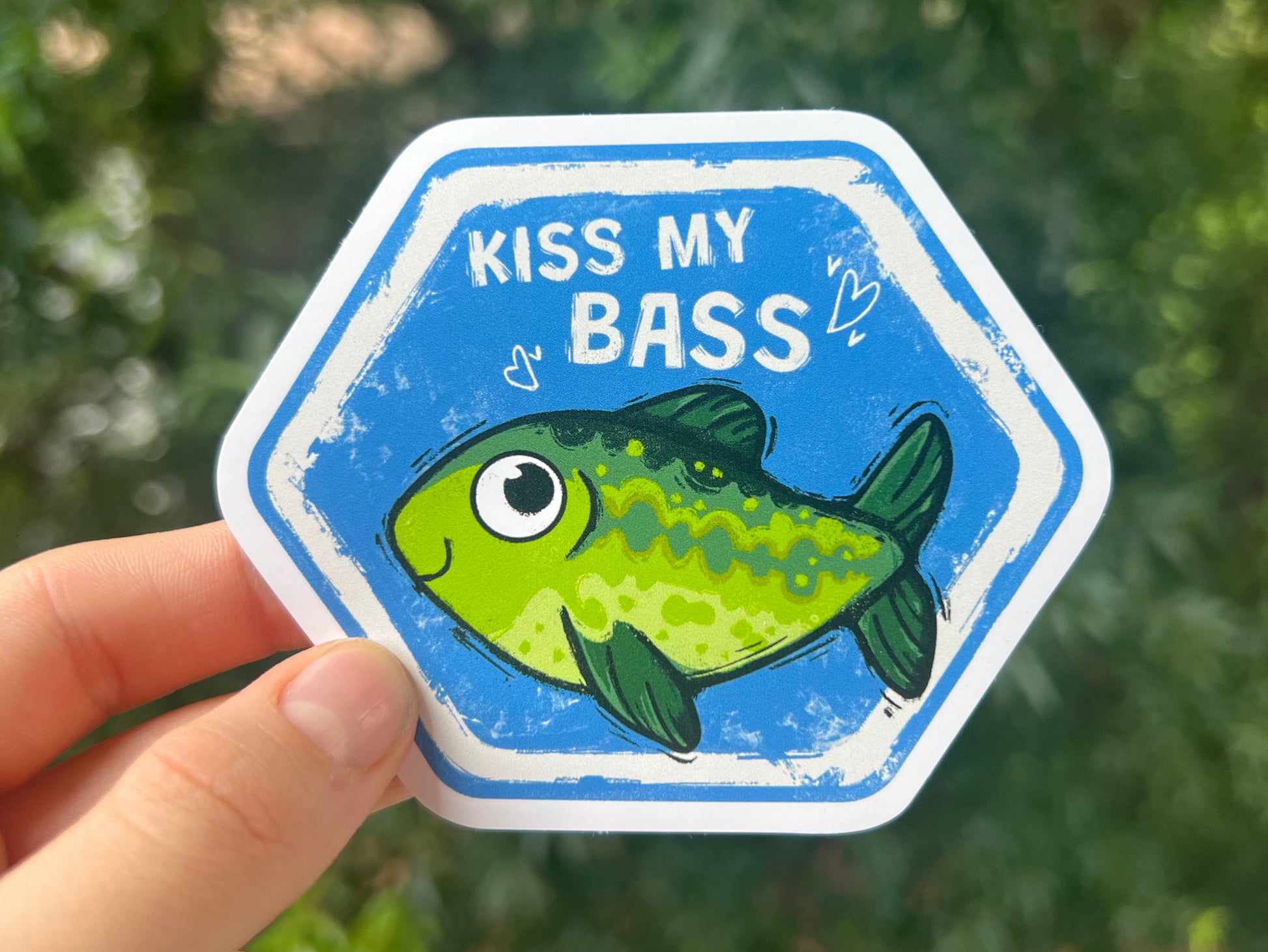 Hexagonal sticker with a fish graphic and text 'Kiss My Bass' held by a hand against a blurred green background.